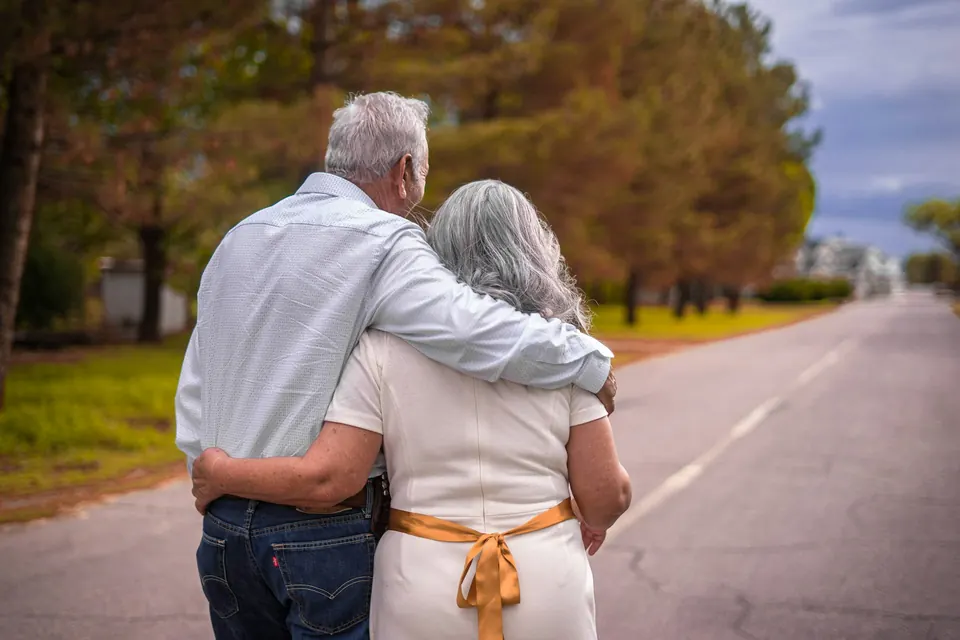 Couple de retraités aux cheveux blancs marchant enlacés sur une route bordée d’arbres, vu de dos